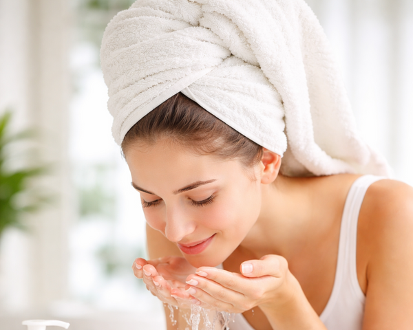 Woman washing her face with a towel on her head, surrounded by skincare products.